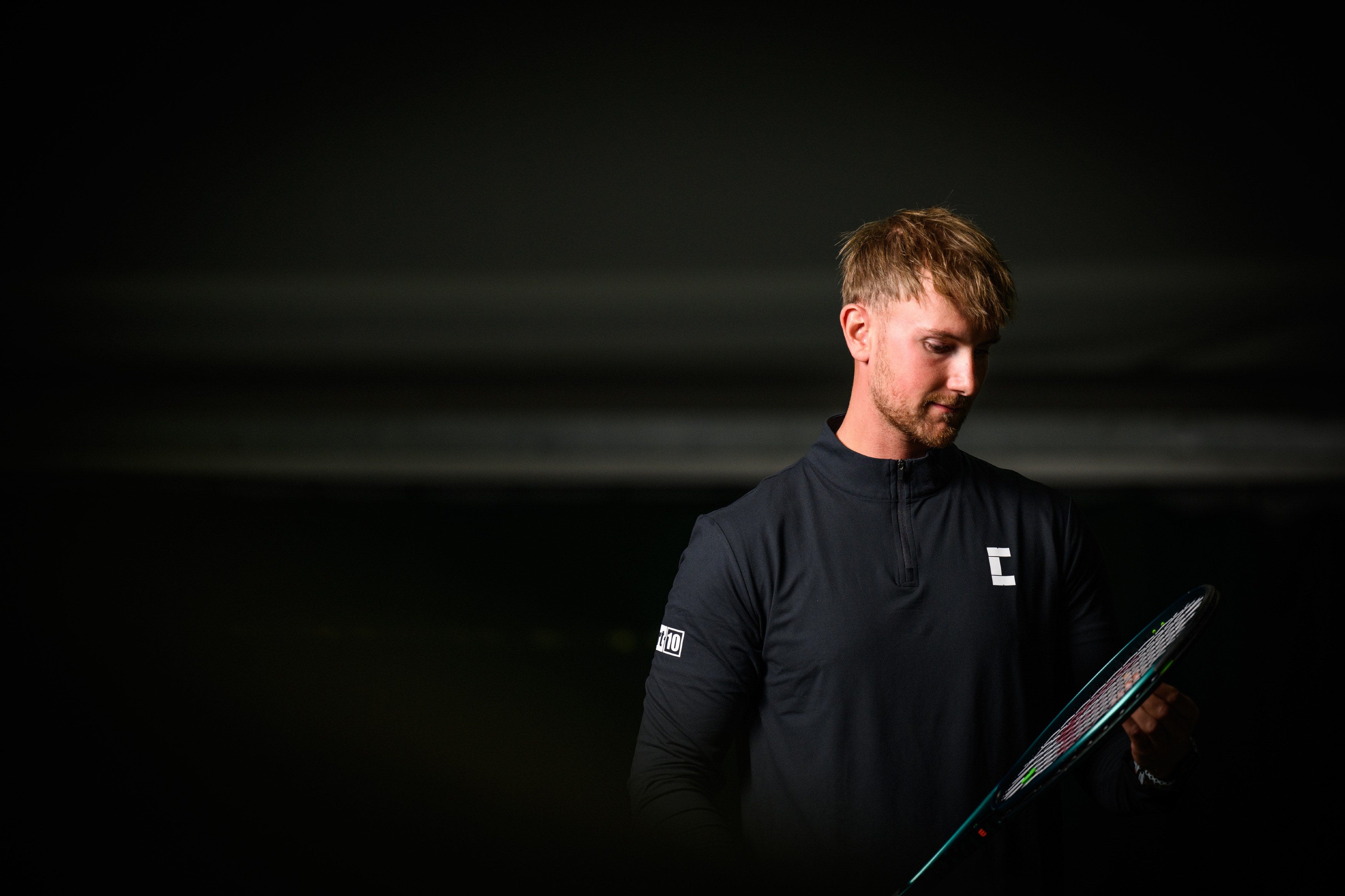 Tennis player holding a tennis racket against a dark background, wearing Court10 Performance Long Sleeve.