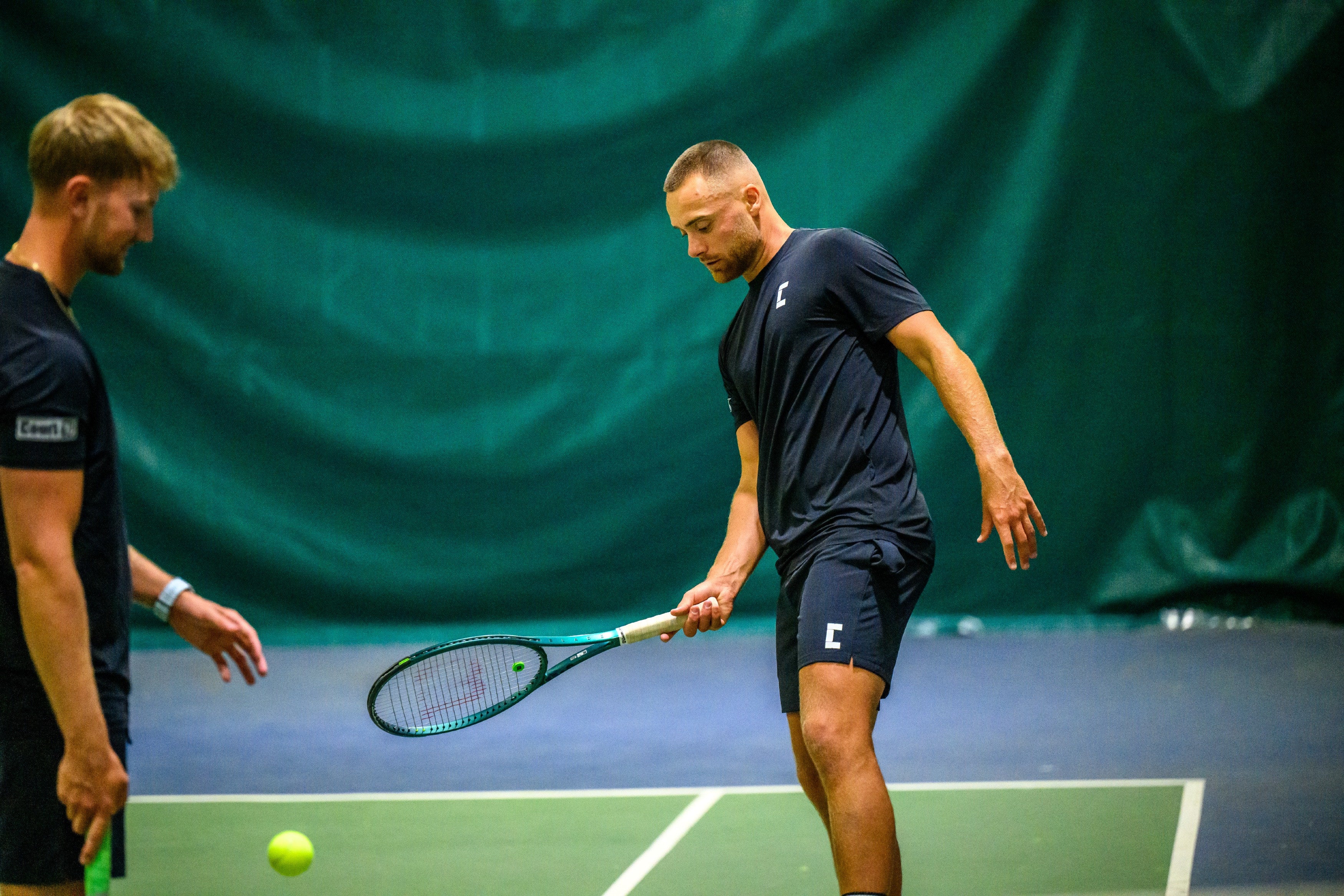 Two men on a tennis court with one preparing to hit a ball, both wearing full Court10 performance apparel.
