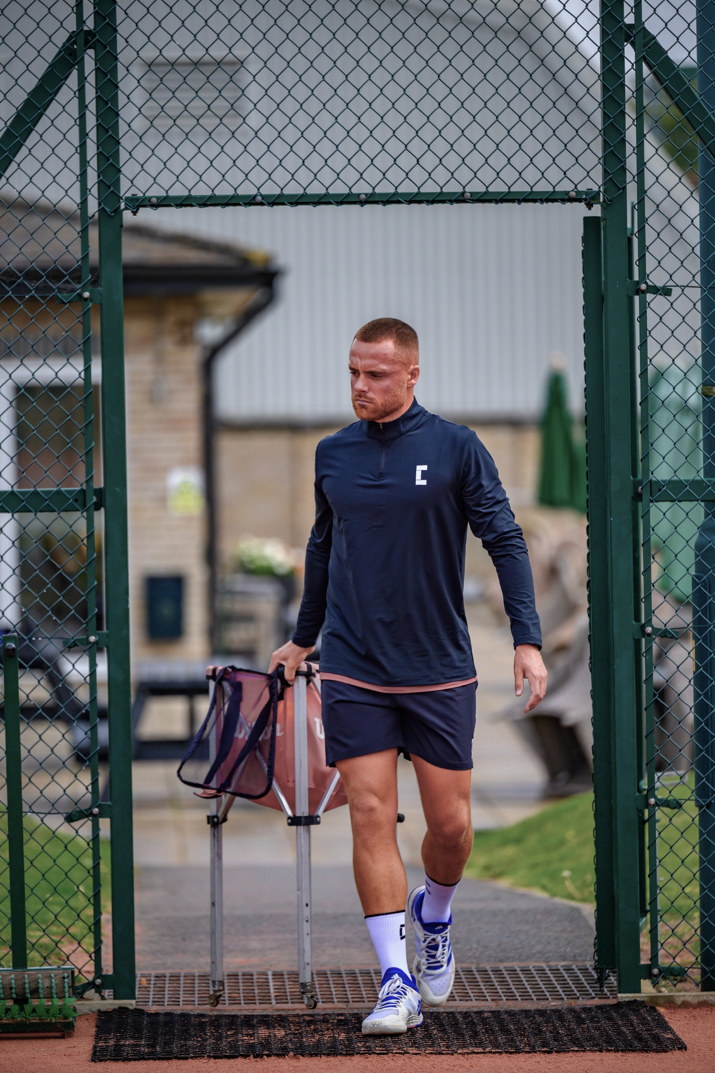 Tennis coach in Court10 performance kit walking through a court gate with sports equipment.